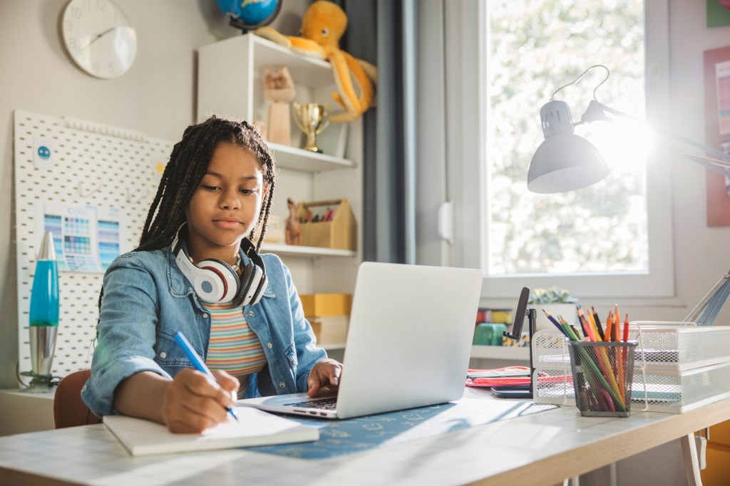 High school student studying in bedroom with laptop, pen and paper, wearing headphones around neck.
