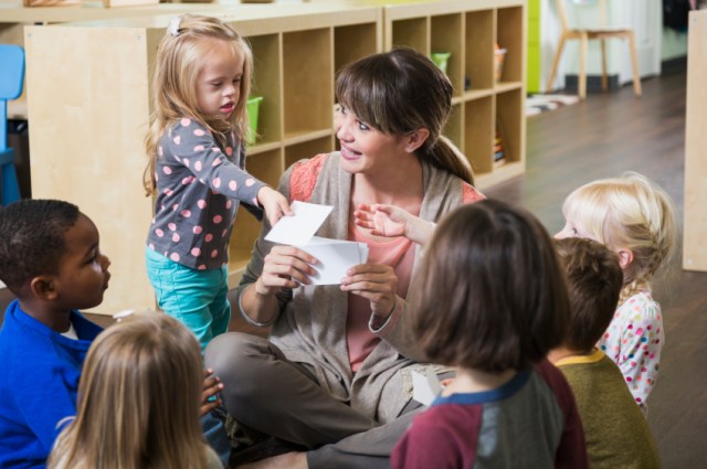 A preschool teacher sitting on the floor with a group of multi-ethnic children in a circle. They are watching her as she holds up a card. A little girl, a special needs child with down syndrome, is standing beside her handing a card to another child.