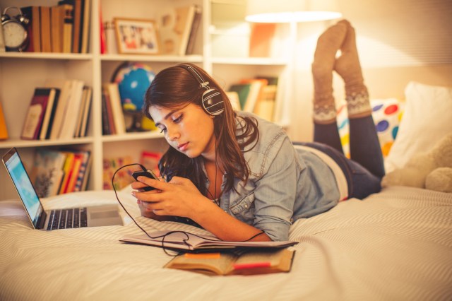 Teen girl on bed, looking at cell phone with headphones on while doing homework