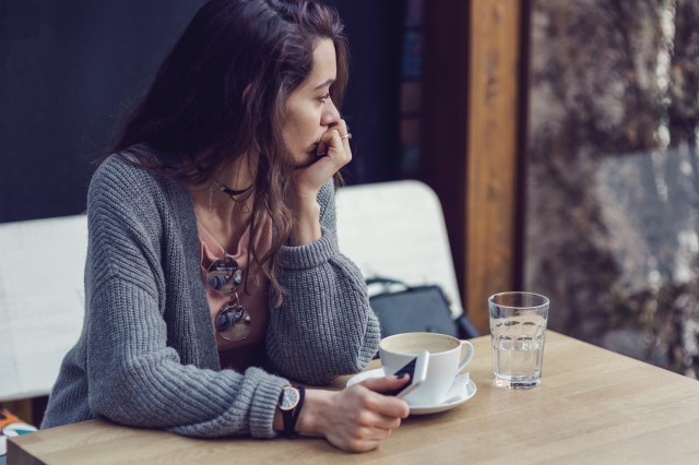 Woman sitting alone, deep in thought, having coffee and holding her mobile phone