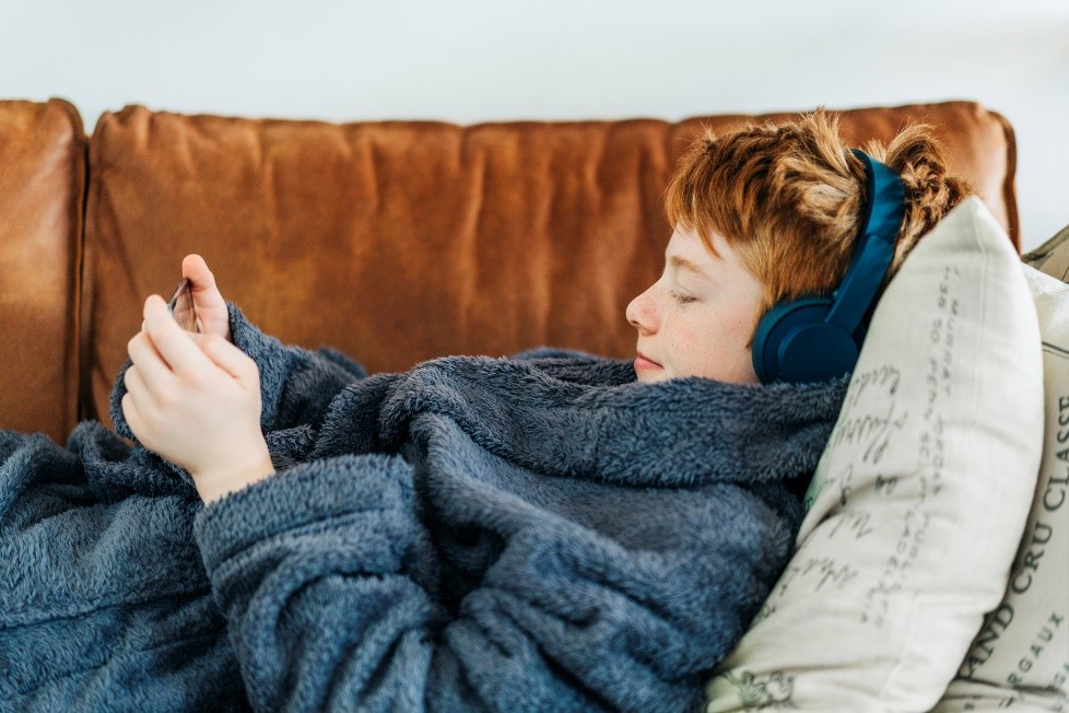 Teen boy in bathrobe lying on couch, wearing headphones and looking at mobile device.