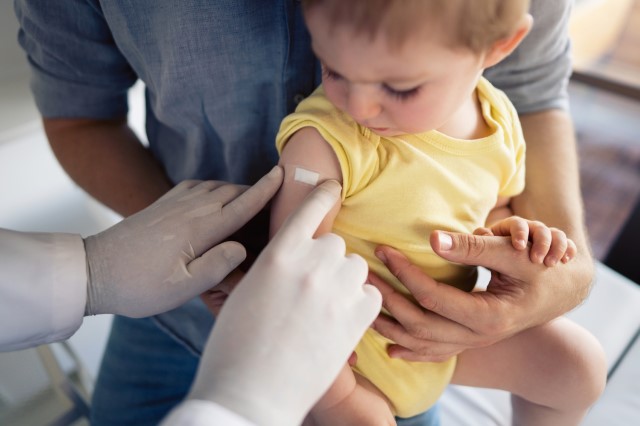 Immunizer's hands affixing bandage to toddler's arm who looks concerned.